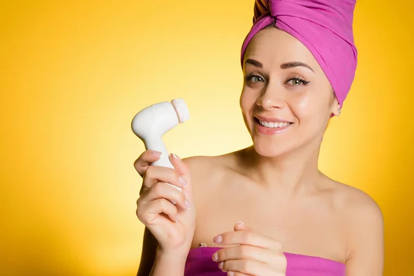 Sorridente bella ragazza con un asciugamano rosa sulla testa facendo una pulizia profonda del viso con una speciale spazzola elettrica — Foto Stock