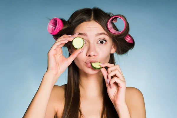 Una divertida chica sorprendida sostiene un trozo de pepino cerca de su cara y se come otro, en la cabeza de su rizador de pelo — Foto de Stock