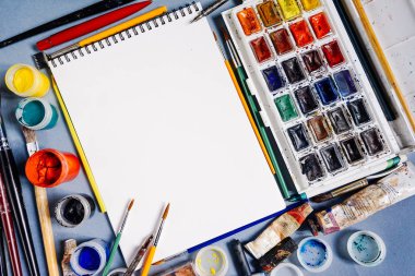 Mavi masa sanatsal aksesuarları, beyaz bir not defteri, suluboya ve guaş, birçok fırçaları vardır