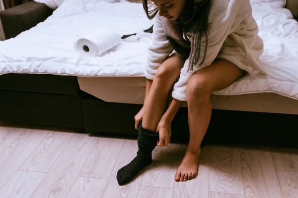 Молодая девушка в белом махровом халате надевает тёплые чёрные носки. Чтобы согреться — стоковое фото