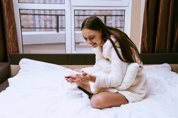 Ένα γέλιο κορίτσι σε μια ρόμπα terry λευκό είναι κάθεται στο κρεβάτι και να τρώνε χυλό βρώμης — Φωτογραφία Αρχείου