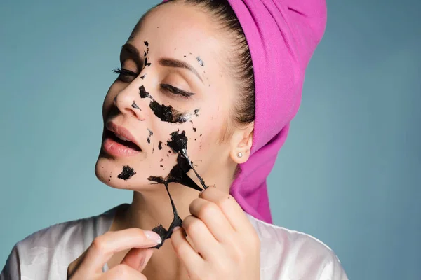 Uma menina atraente com uma toalha rosa na cabeça remove a máscara de limpeza preta de seu rosto — Fotografia de Stock