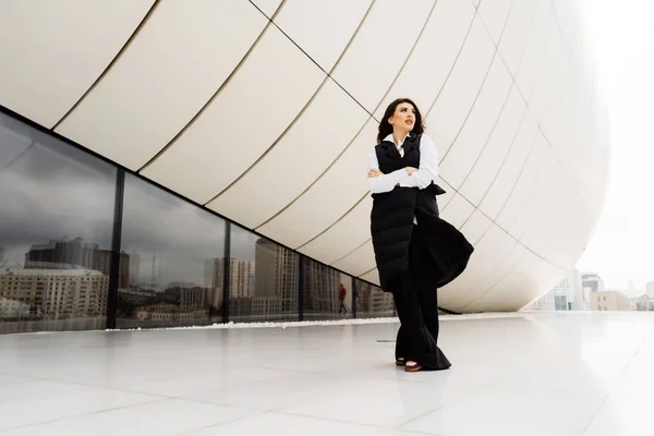 Mujer con estilo en un traje negro y camisa blanca posando sobre el fondo de un edificio moderno inusual en la ciudad de Bakú — Foto de Stock