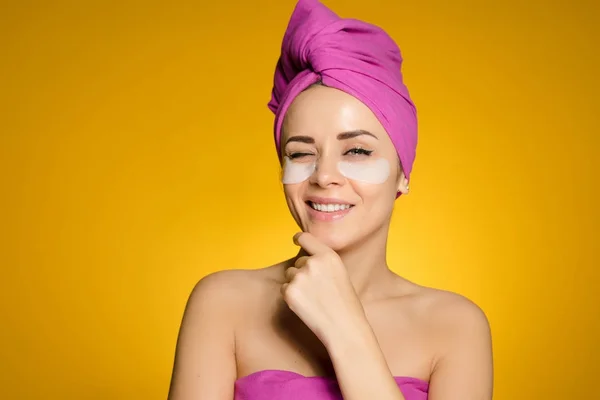 Χαμογελώντας ελκυστικό κορίτσι με μια ροζ πετσέτα στο κεφάλι της, κάτω από τα μάτια των patches με σιλικόνη — Φωτογραφία Αρχείου