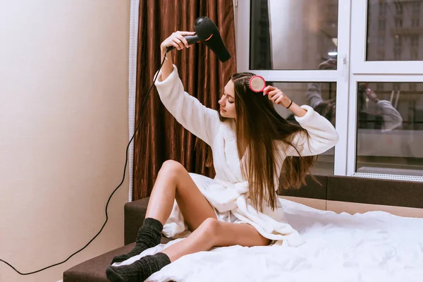 Молодая девушка в белом махровом халате сидит на кровати и сушит длинные волосы феном — стоковое фото