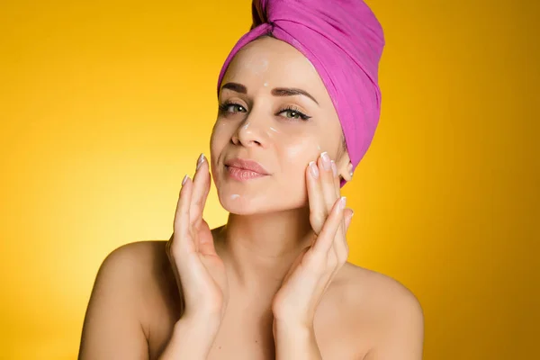 Ελκυστική κοπέλα με μια ροζ πετσέτα στο κεφάλι της, βάζει μια ενυδατική κρέμα στο πρόσωπό της — Φωτογραφία Αρχείου