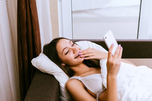 Красивая сонная девушка лежит в постели под одеялом, зевает и смотрит на свой смартфон — стоковое фото
