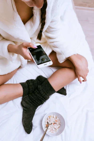 Een meisje in een badjas wit terry en warme sokken is zittend op het bed, eten van havermoutpap, een plaat fotograferen — Stockfoto