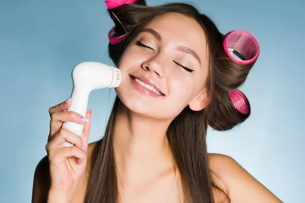 Χαμογελώντας όμορφο κορίτσι να κάνει ένα βαθύ καθαρισμό του προσώπου με ειδική ηλεκτρική βούρτσα, στο κεφάλι του το ψαλίδι για μπούκλες — Φωτογραφία Αρχείου