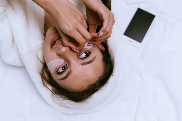 Bela menina atraente encontra-se na cama, cola adesivos de silicone sob os olhos, ao lado do telefone encontra-se — Fotografia de Stock