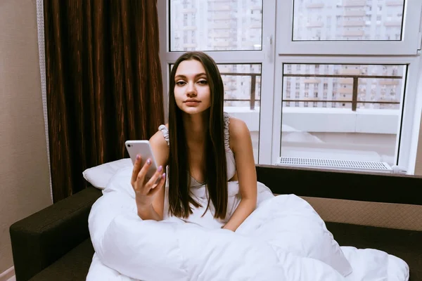 Длинноволосая девушка только что проснулась, сидит в постели под белым одеялом, держит в руке смартфон. — стоковое фото