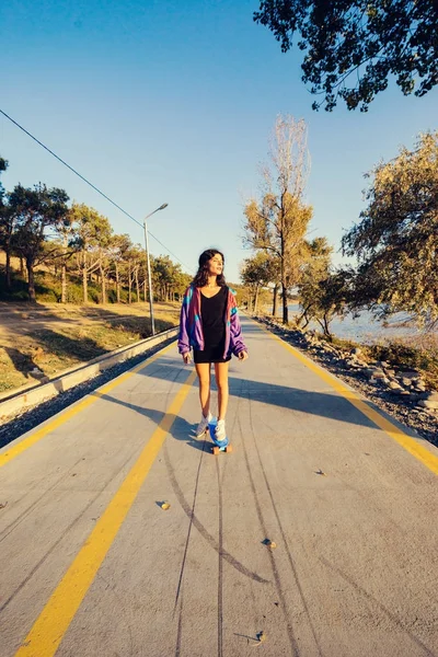 Una joven delgada con estilo en una chaqueta púrpura patina en un monopatín en la carretera, en los rayos del sol — Foto de Stock
