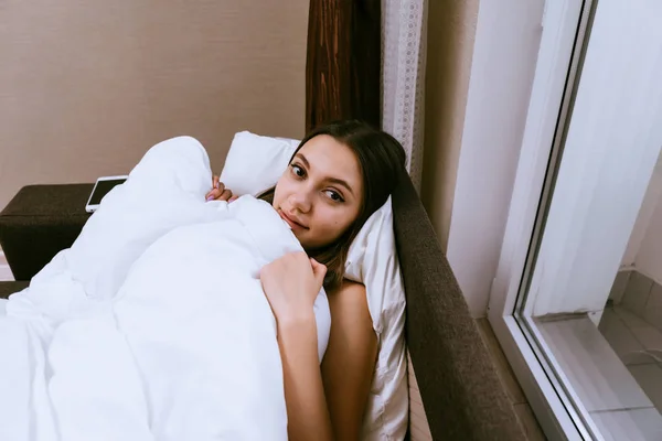 Csinos fiatal lány fekszik az ágyban, a fehér takaró alatt, nem akar-hoz kelj fel — Stock Fotó