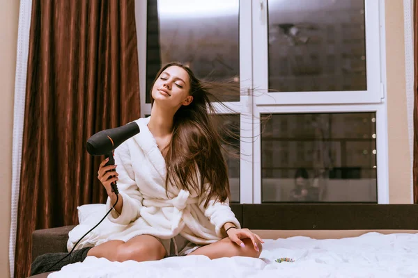 Привлекательная молодая девушка в белом махровом халате сидит на кровати и сушит длинные волосы феном — стоковое фото