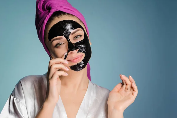 Menina sorridente atraente com uma toalha rosa na cabeça remove uma máscara preta de seu rosto contra pontos pretos — Fotografia de Stock