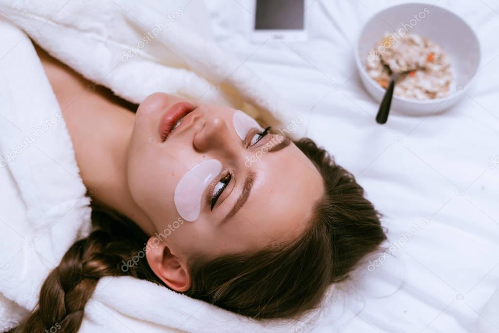 young girl lying on a bed in a white coat, under the eyes of silicone patches against swelling, next to a plate of oatmeal porridge