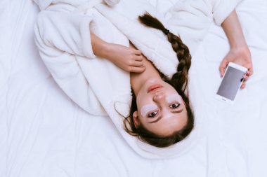 güzel bir genç kız erken, beyaz bir ceket, bir akıllı telefon elinde tutan bir yatak, silikon yamalar, gözleri altında yatan uyandım