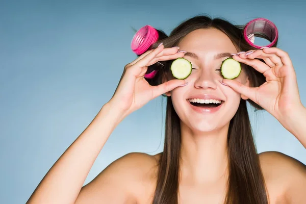 Happy usmívající se dívka drží kus okurky v rukou hydratují pokožku na tváři, na její hlavě natáčky — Stock fotografie