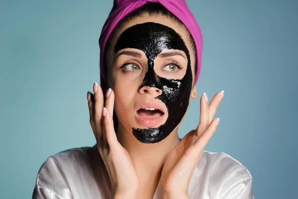 Piękna młoda kobieta z różowy ręcznik na głowie dotyczy czarna maska oczyszczenie przed czarnymi kropkami, wygląda zaskoczony — Zdjęcie stockowe