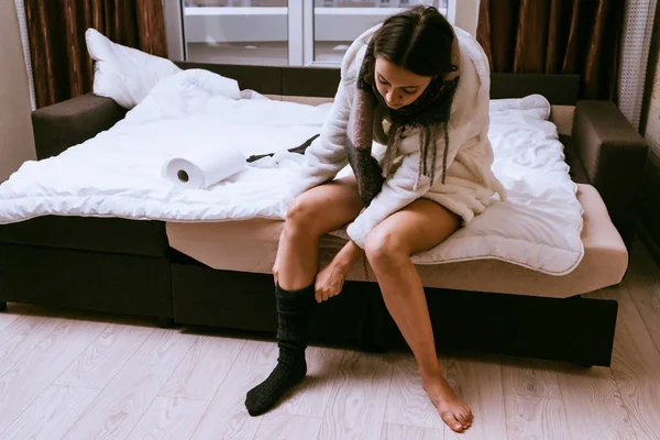 Uma jovem menina em um vestido de vestir branco terry senta-se na cama, adoece, coloca meias quentes — Fotografia de Stock