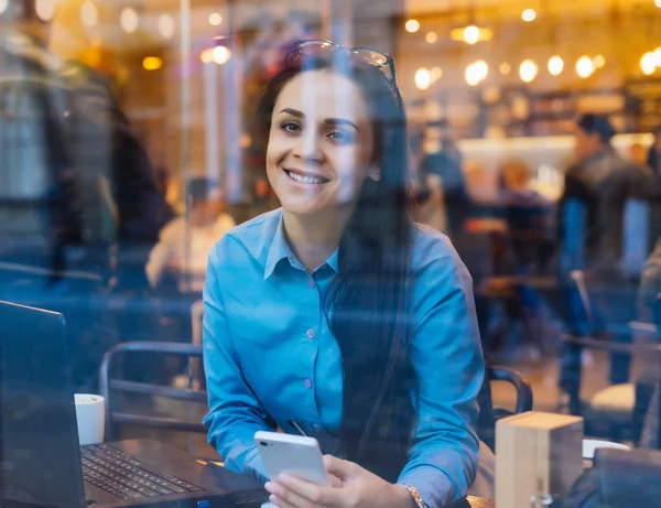 Feliz jovem menina freelance bem sucedido sentado no café pela janela, trabalhando no laptop e smartphone — Fotografia de Stock