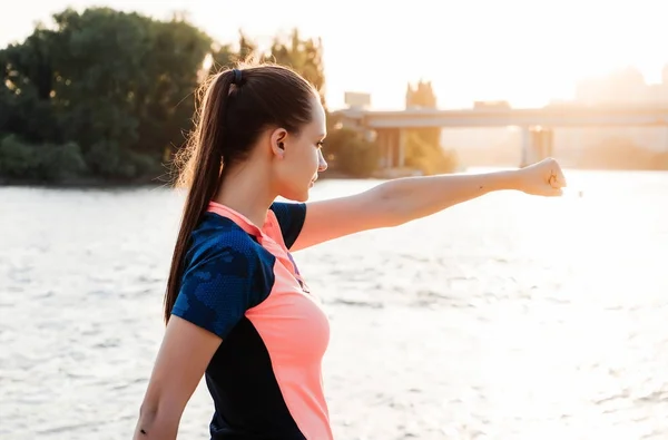 Långhåriga flicka i sportkläder knådar muskler mot bakgrund av floden och sunset — Stockfoto