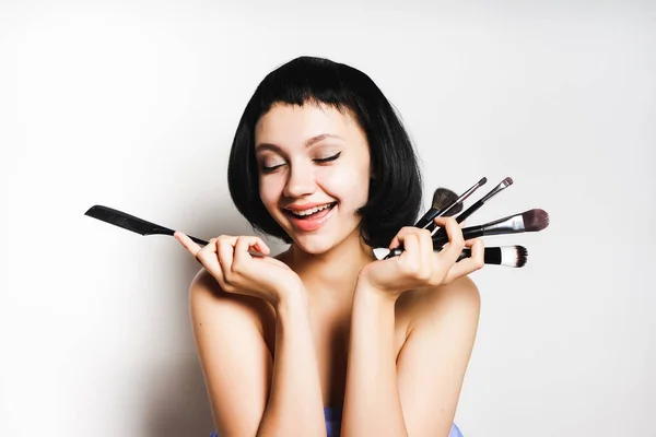 Menina sorridente feliz com cabelo preto curto está segurando escovas de maquiagem cosmética nas mãos — Fotografia de Stock