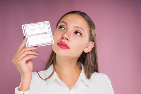 एक सफेद शर्ट में एक युवा सुंदर लड़की झूठी eyelashes के साथ एक लपेटा हुआ पैकेज पकड़ रही है, ध्यान से ऊपर की ओर देख रही है — स्टॉक फ़ोटो, इमेज
