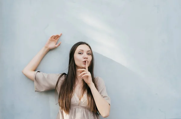 Attraente giovane ragazza modello ha messo un dito alle labbra, è in posa da un muro leggero — Foto Stock