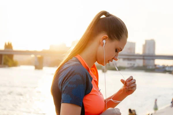 Vacker ung flicka i sportkläder som lyssnar på musik i hörlurar på bakgrunden av floden och solen — Stockfoto