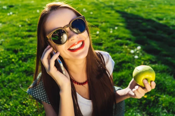 Улыбающаяся женщина разговаривает с телефоном, держит зеленое яблоко, сидит на зеленой траве в парке — стоковое фото