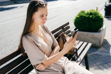 bej renkli elbiseli çekici güzel kız modeli Güneş ışınlarının ahşap bir bankta oturur, bir akıllı telefon tutar