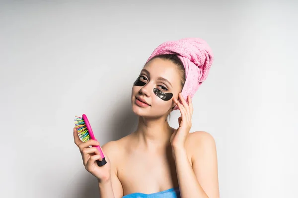 一个年轻漂亮的女孩照顾自己, 在她的头上, 粉红色的毛巾, 在黑色硅胶片的釉下, 在她手中拿着一把梳子 — 图库照片