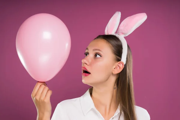 一个年轻的女孩看起来惊讶于一个粉红色的空气气球, 在她的头上的兔子耳朵的形式边缘 — 图库照片