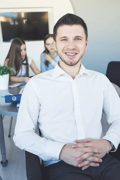 Молодой уверенный в себе успешный мужчина, офисный работник, в белой рубашке улыбается, на заднем плане две девушки работают — стоковое фото