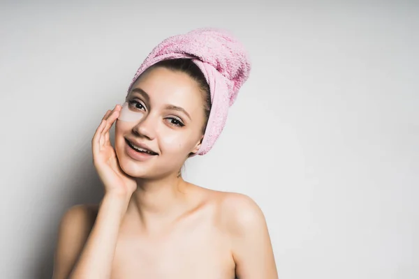 Sorrindo menina radiante cuida de si mesma, na cabeça uma toalha rosa, sob os olhos de adesivos hidratantes de silicone — Fotografia de Stock