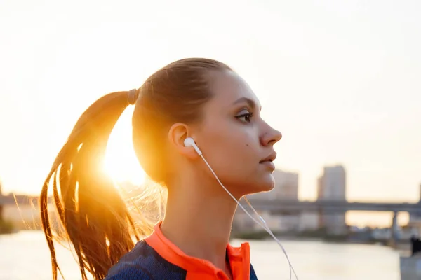 young purposeful girl runs by the river at sunset, listens to music on headphones