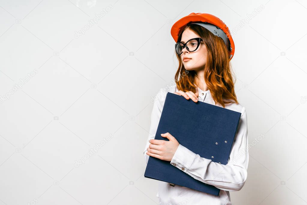 confident young red-haired girl builder wearing an orange protective helmet and wearing glasses keeps project documentation in hand
