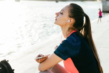çekici bir genç kız spor içinde eğitim, kulaklık müzik dinlemek, nehirde seyir sonra dinleniyor
