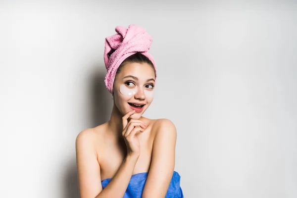 Ελκυστικό κορίτσι λάμπει με μια ροζ πετσέτα στο κεφάλι της, έξυπνα χαμόγελα, κάτω από τα μάτια των patches με σιλικόνη — Φωτογραφία Αρχείου