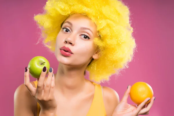 Молодая смешная девушка с зеленым яблоком и оранжевым, в жёлтом клоунском парике — стоковое фото