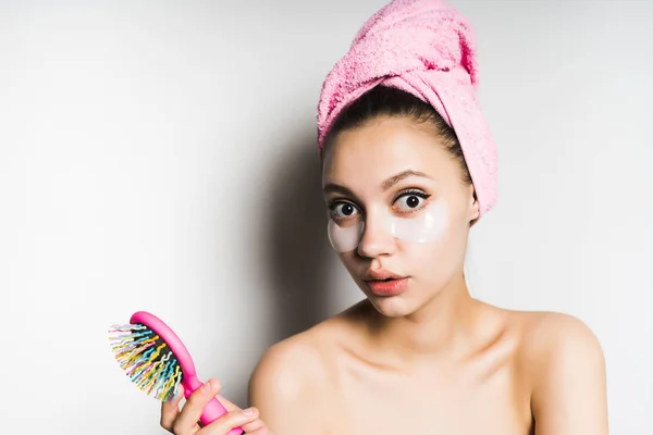 Sorprendida joven hermosa chica cuida de sí misma, en su cabeza una toalla rosa, bajo los ojos de parches de silicona, sostiene un peine en su mano — Foto de Stock