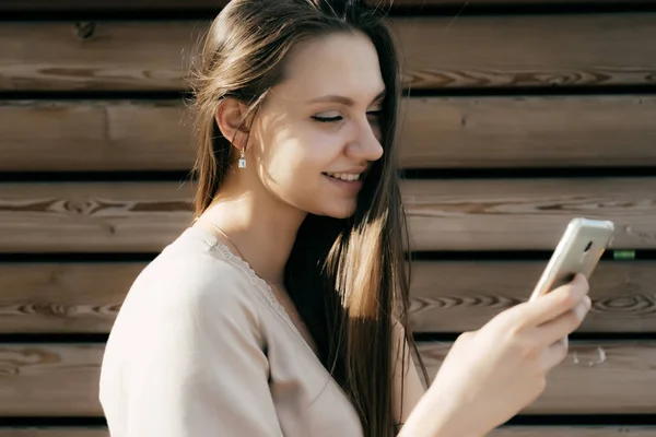 Felice ragazza sorridente in abito beige guardando nel suo smartphone, su uno sfondo di legno — Foto Stock