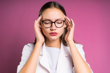 Gözlüklü genç bir kız kafasını acıyor, gözleri kapalı, pembe bir arka plan