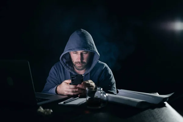 Homme intelligent pirate assis dans le noir à l'ordinateur portable, tenant un smartphone dans ses mains, piratage quelque chose — Photo