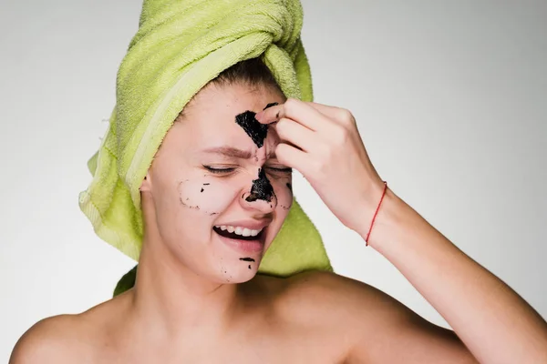 Una joven con una toalla verde en la cabeza se quita una máscara de limpieza negro contra puntos negros, sensaciones desagradables — Foto de Stock