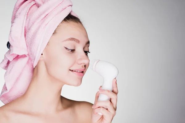 Souriant jeune fille avec une serviette rose sur la tête faire un nettoyage en profondeur de la peau sur son visage avec une brosse électrique — Photo