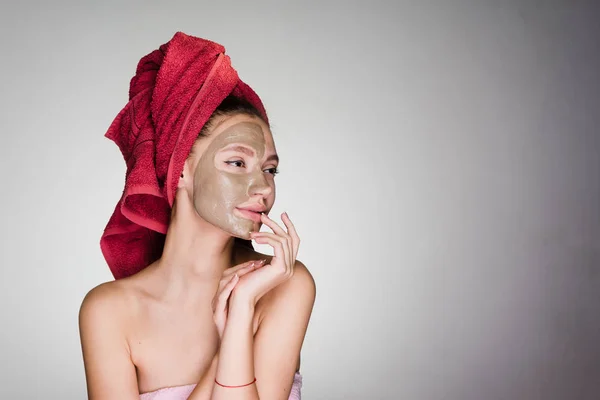 Gyönyörű lány gondoskodik a magát, a spa szolgáltatásait, agyagos maszk arcán, a fején egy piros törölköző — Stock Fotó