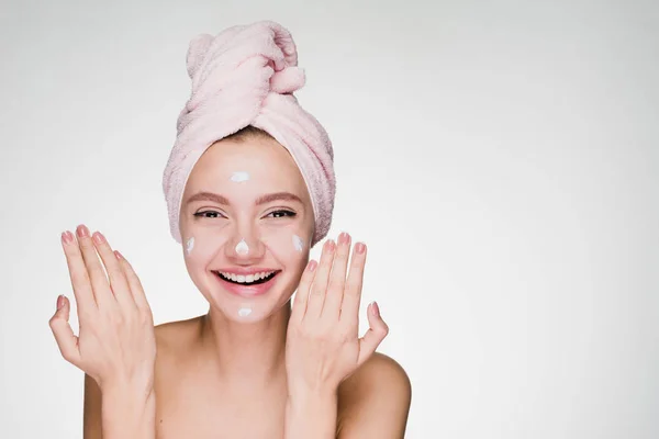 Счастливая молодая девушка хочет хорошо выглядеть, смеется, на голове розовое полотенце, нанес белый крем на ее лицо — стоковое фото