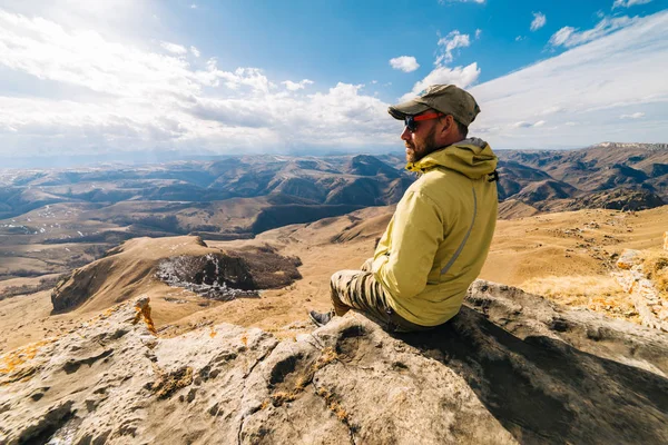 A male traveler in sunglasses sits on the edge of a cliff, enjoying mountains and clear air — Stock Photo, Image